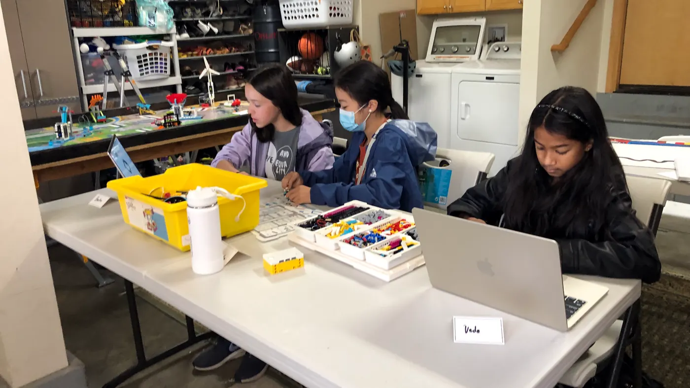 Three Advanced class students at a table, two building from LEGO parts trays and one coding on a laptop