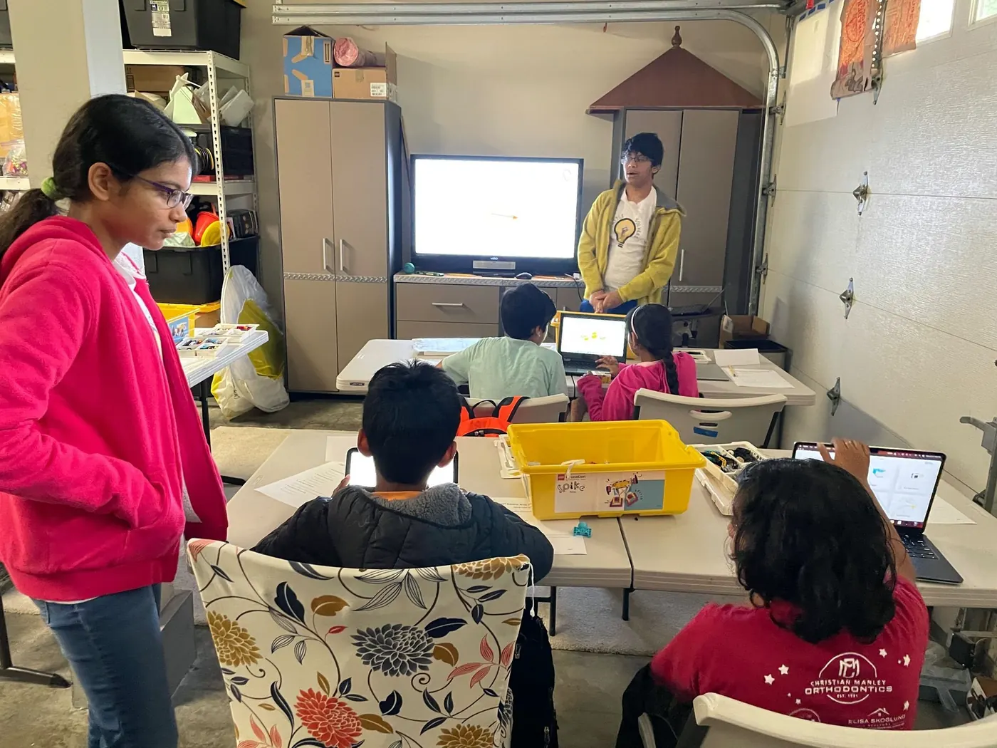 Students at laptops around a LEGO bin while a team member presents on a TV