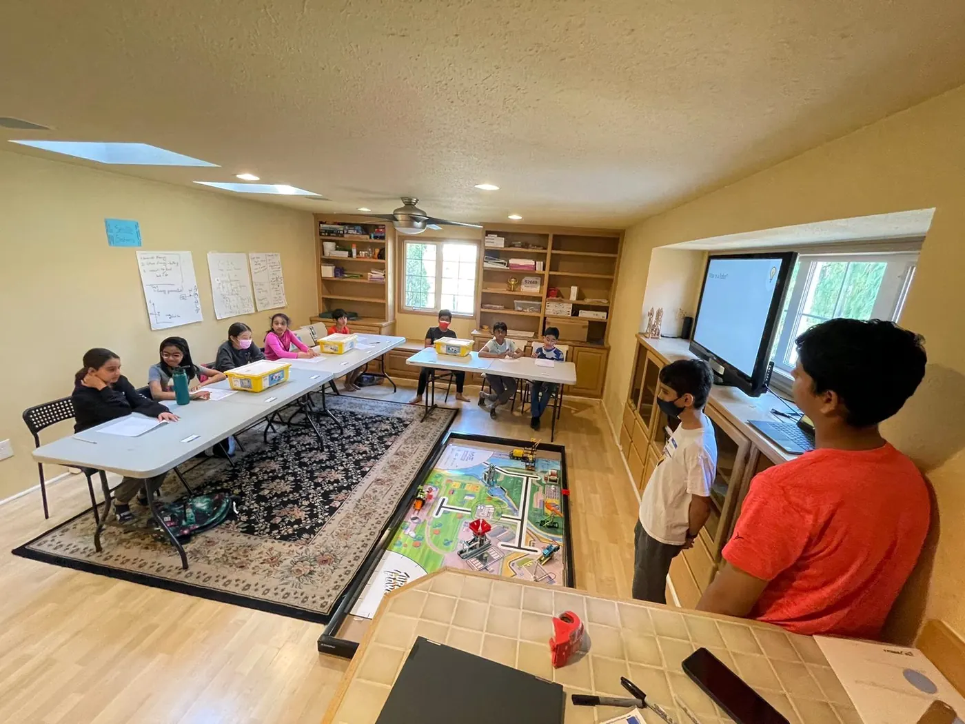A classroom of students at two long tables, with the FIRST LEGO League mat on the floor and a team member at the screen