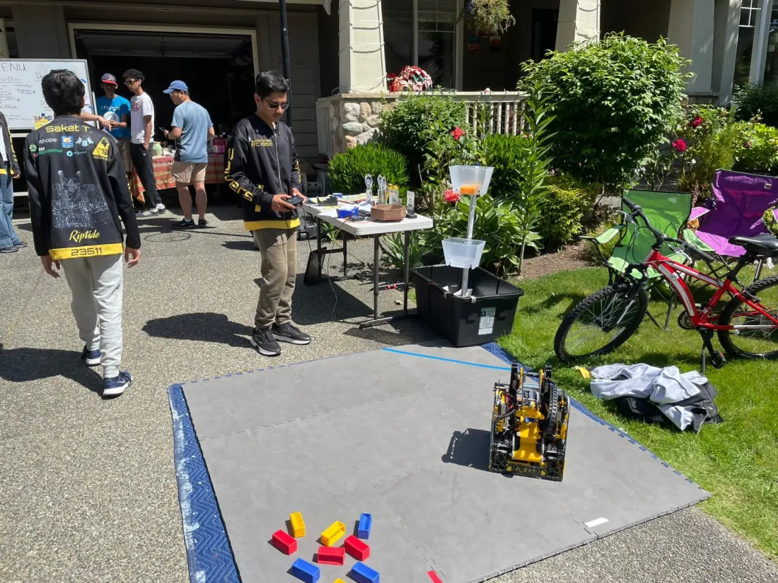 The robot on a foam-tile demo field set up on a driveway, with team members and sale tables behind it