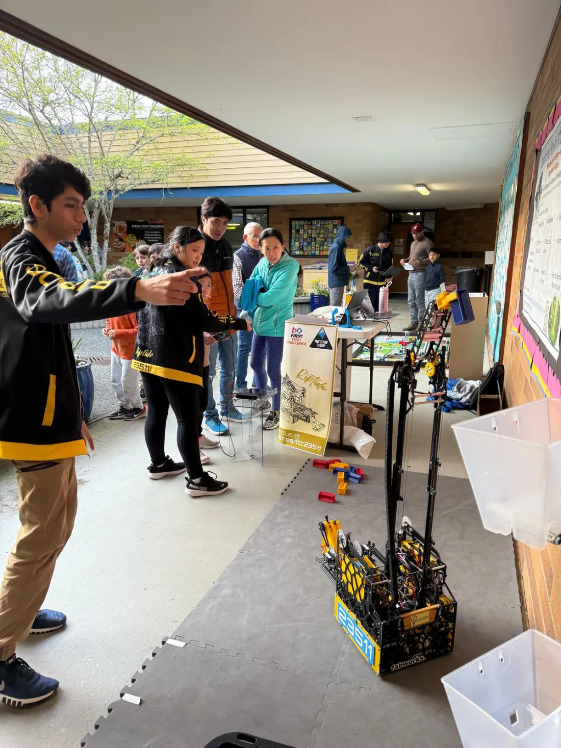 A team member points to the robot while students and parents look on in a covered school walkway