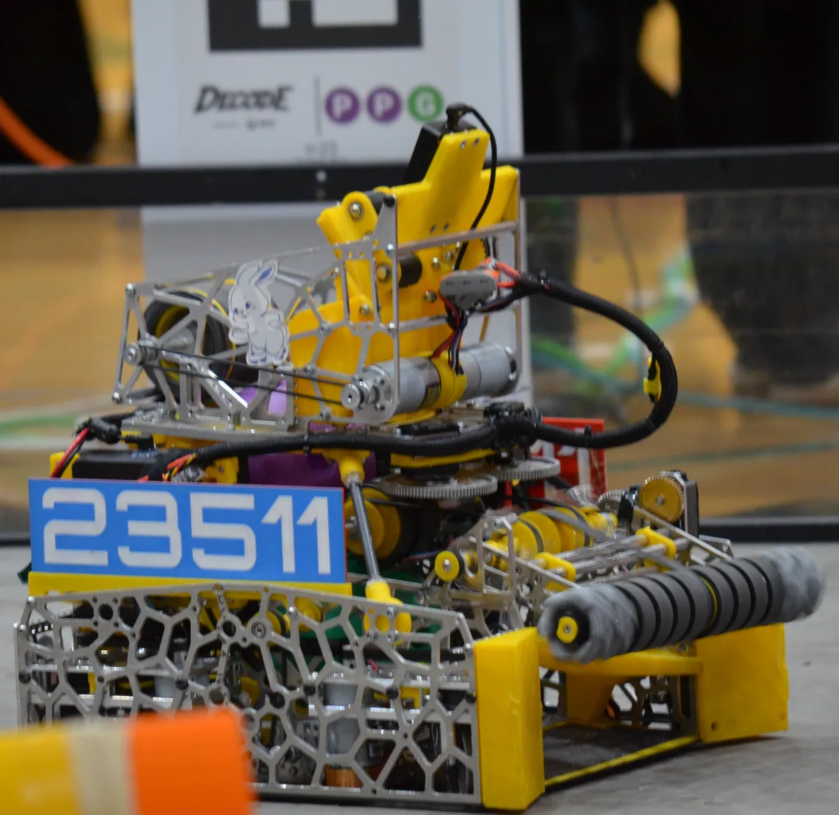 Cypher v1 on a DECODE field: a yellow turret and launcher above the swerve chassis, a blue 23511 plate on the front and the pivoting intake with gray rollers at right
