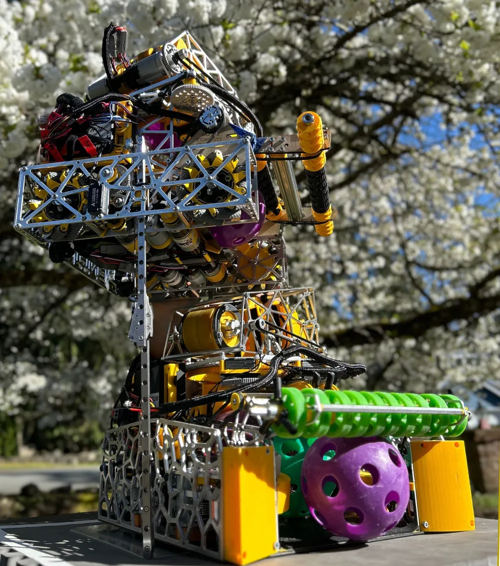 Enigma raised on its linear-rail lift outdoors under a flowering cherry tree: a silver truss frame and yellow gears up top, a green intake roller and a purple ball at its base