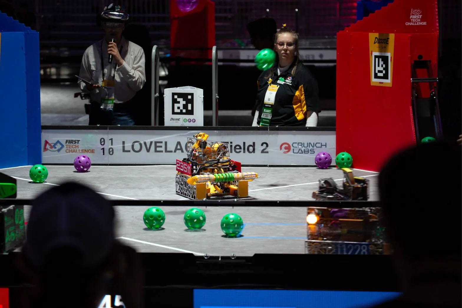Cypher, with a red 23511 alliance sign and its green roller intake, on Lovelace Field 2 at the FIRST World Championship, green and purple balls on the tiles, and an announcer and a volunteer behind the wall