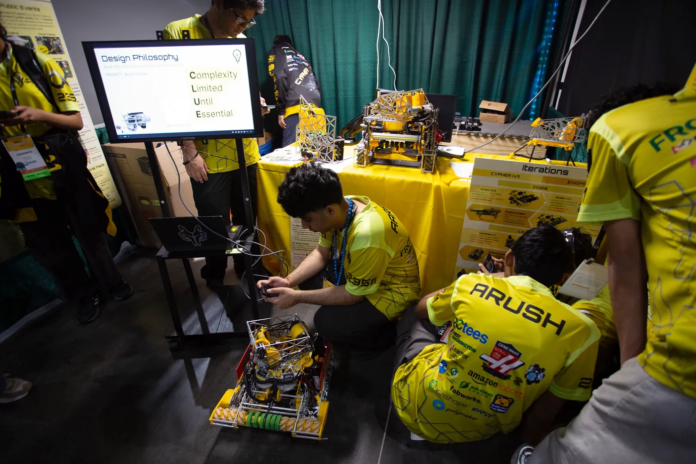 Team members in yellow jerseys working on Cypher on the floor of their Houston pit, a second robot on the table behind them, beside a Design Philosophy slide and an Iterations board